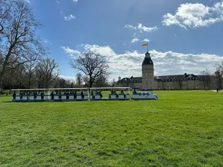 Die Karlsruher Schlossgartenbahn mit der blau-weißen Porsche-Lok bei ihrer Fahrt durch den Schlossgarten mit dem Schloss im Bildhintergrund.