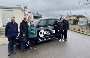 Das Foto zeigt (von links nach rechts) Taxiunternehmer Dirk Holl, Olaf Strotkötter (Prokurist KVV), Bürgermeisterin Nicola Bodner, Bürgermeister Timur Özcan und Ragnar Watteroth(Finanzdezernent Landkreis Karlsruhe), die vor einem MyShuttle-Fahrzeug stehen. Im Hintergrund hält eine AVG-Stadtbahn.