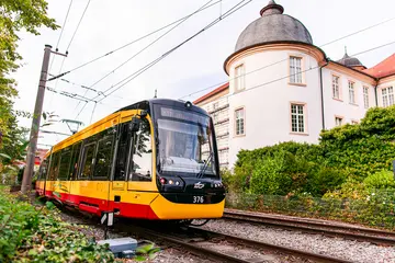 Eine gelb-rote Straßenbahn fährt an dem Schloss in Ettlingen vorbei. Der Himmel ist leicht bewölkt.