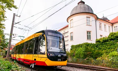 Eine gelb-rote Straßenbahn fährt an dem Schloss in Ettlingen vorbei. Der Himmel ist leicht bewölkt.