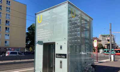 Ein Aufzug des Karlsruher Stadtbahntunnels am Durlacher Tor. An das Munhaus des Aufzuges ist ein Fahrrad angelehnt. Im Hintergrund sind Häuserfassaden, eine Trambahn und der blaue Himmel zu sehen.