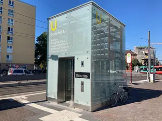 Ein Aufzug des Karlsruher Stadtbahntunnels am Durlacher Tor. An das Munhaus des Aufzuges ist ein Fahrrad angelehnt. Im Hintergrund sind Häuserfassaden, eine Trambahn und der blaue Himmel zu sehen.