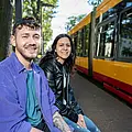 Eine junge Frau und ein junger Mann sitzen an einer Haltestelle im Grünen. Im Hintergrund sieht man die dahliengelb-rote Bahn. 