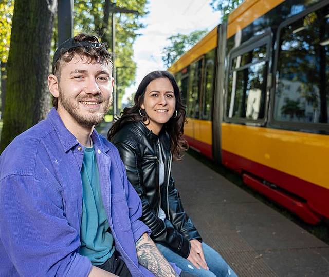 Eine junge Frau und ein junger Mann sitzen an einer Haltestelle im Grünen. Im Hintergrund sieht man die dahliengelb-rote Bahn.