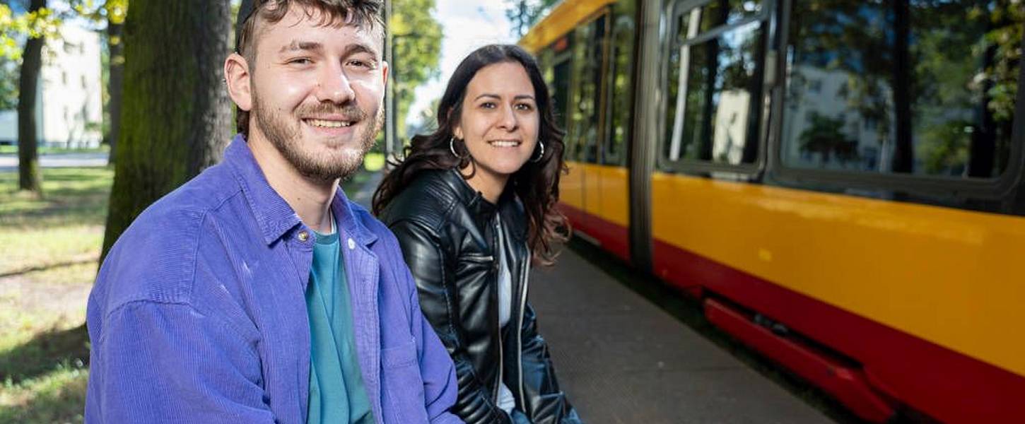 Eine junge Frau und ein junger Mann sitzen an einer Haltestelle im Grünen. Im Hintergrund sieht man die dahliengelb-rote Bahn.