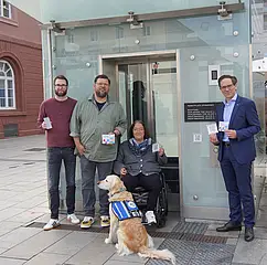 Vor dem Stadtbahn-Tunnel am Marktplatz sind von rechts nach links folgende Personen zu sehen: Christian Höglmeier (technischer Geschäftsführer der VBK), Ulrike Wernert (kommunale Behindertenbeauftragte der Stadt Karlsruhe), Artur Bundnik (Projektstelle Web-App „Karlsruhe barrierefrei“) und Marcus Albert (System-Administrator „Karlsruhe barrierefrei“). Sie halten die Aufkleber mit den QUR-Codes zu "Karlsruhe barrierefrei" in der Hand. Vor ihnen sitzt ein Hund. 