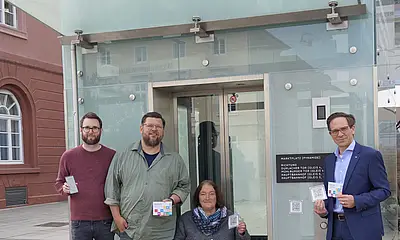 Vor dem Stadtbahn-Tunnel am Marktplatz sind von links nach rechts folgende Personen zu sehen: Christian Höglmeier (technischer Geschäftsführer der VBK), Ulrike Wernert (kommunale Behindertenbeauftragte der Stadt Karlsruhe), Artur Bundnik (Projektstelle Web-App „Karlsruhe barrierefrei“) und Marcus Albert (System-Administrator „Karlsruhe barrierefrei“). Sie halten die Aufkleber mit den QUR-Codes zu "Karlsruhe barrierefrei" in der Hand. Vor ihnen sitzt ein Hund. 