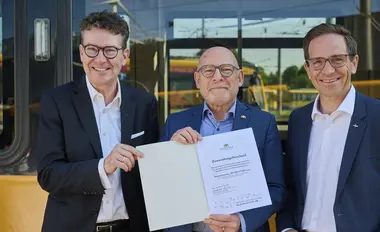 Landes-Verkehrsminister Winfried Hermann (Mitte) steht vor einer Trambahn und hält den Zuwendungsbescheid für die Verkehrsbetriebe Karlsruhe in der Hand. Neben ihm stehen Prof. Dr. Alexander Pischon (links) und Christian Höglmeier (rechts) von der VBK-Geschäftsleitung.