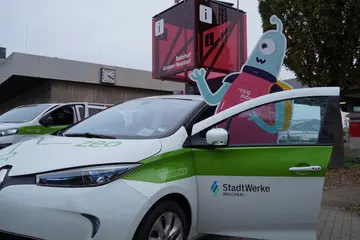 Ein Elektroauto des Car-Sharing-Anbieters ZEO steht vor dem regiomove-Port am Bahnhof in Graben-Neudorf. Aus der geöffneten Fahrertür guckt ein Alien aus Pappe heraus. Dieser steht für das regiomove-Motto "Alles außer beamen."