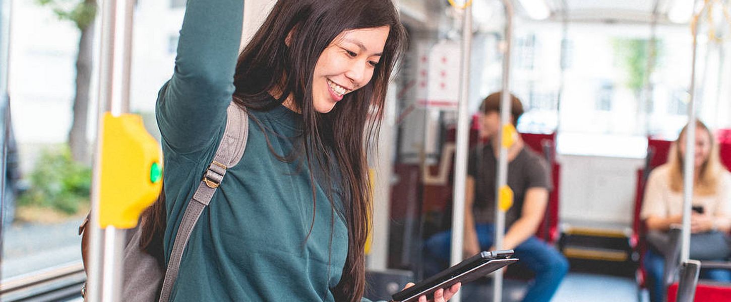 Eine Frau steht lächelnd in einer Bahn. Sie schaut auf ein Tablet in ihrer Hand.