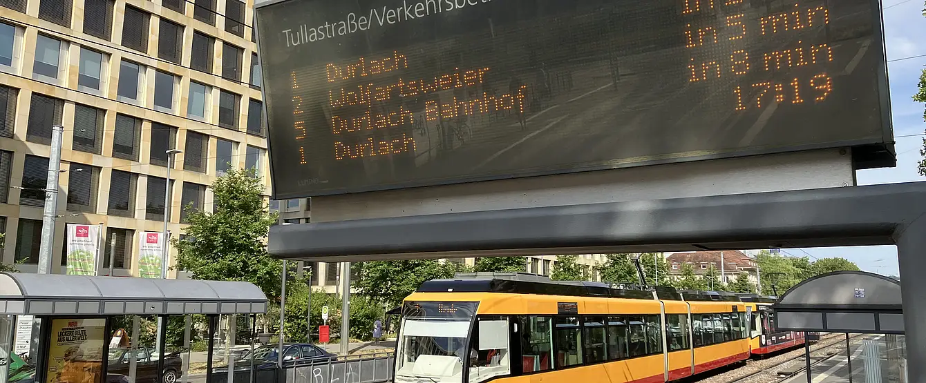 Eine Trambahn der Verkehrsbetriebe Karlsruhe fährt in die Haltestelle Tullastraße/Alter Schalchthof ein. Im Bildvordergrund ist ein Haltestellen-Anzeiger für die Fahrgastinformation zu sehen.