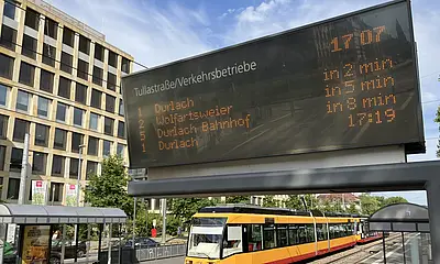 Eine Trambahn der Verkehrsbetriebe Karlsruhe fährt in die Haltestelle Tullastraße/Alter Schalchthof ein. Im Bildvordergrund ist ein Haltestellen-Anzeiger für die Fahrgastinformation zu sehen.