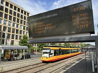 Eine Trambahn der Verkerhsbetriebe Karlsruhe fährt in eine Haltestelle ein. Im Vordergrund ist ein Haltestellen-Anzeiger zur Fahrgastinformation zu sehen.