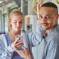 Eine junge Frau und ein junger Mann stehen in einer Bahn. Beide halten ein Smartphone in der Hand.Die Frau schaut auf ihr Smartphone. Der Mann geradeaus und lächelt.