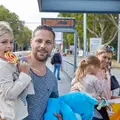 Eine Familie steht an einer Bahnhaltestelle. Der Mann hält ein Mädchen mit Lolli auf dem Arm. Die Frau steht vor dem Fahrkartenautomat. Sie hält ebenfalls ein Mädchen auf dem Arm. Das Mädchen hat einen Fahrschein in der Hand.