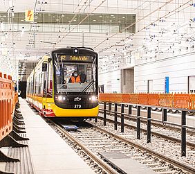 Einfahrende Bahn in einer Haltestelle des neuen Stadtbahntunnels. Rechts im Bild stehen orangefarbene Absperrgitter aus Kunststoff.