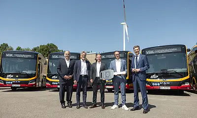 Vor einem Bus steht eine Gruppe von Männern. Im Hintergrund ist ein Windrad zu sehen. 