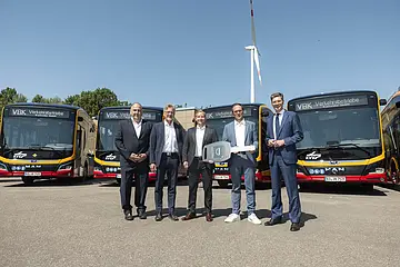 Vor einem Bus steht eine Gruppe von Männern. Im Hintergrund ist ein Windrad zu sehen. 