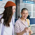 Zwei junge Frauen stehen in einem Bahnhof und blicken strahlend auf ein Smartphone, das eine der beiden Frauen in der Hand hält.