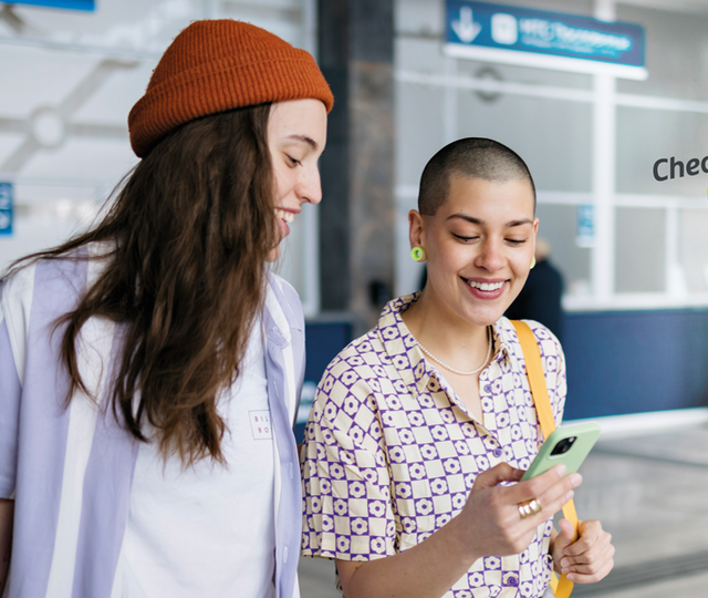 Zwei junge Frauen stehen in einem Bahnhof und blicken strahlend auf ein Smartphone, das eine der beiden Frauen in der Hand hält.