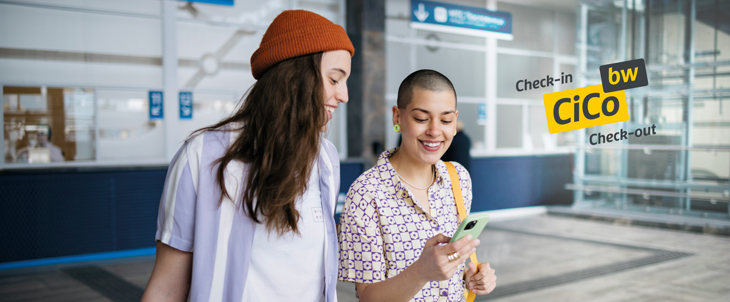 Zwei junge Frauen stehen in einem Bahnhof und blicken strahlend auf ein Smartphone, das eine der beiden Frauen in der Hand hält.