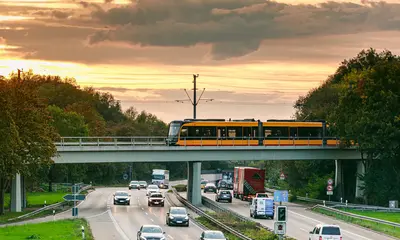 Eine Bahn der AVG überquert auf einer Brücke die Südtangente. Auf der Südtangente fahren Autos. Der Himmel ist bewölkt.