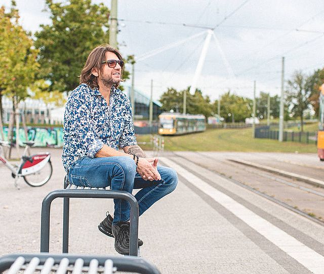 Ein Mann mittleren Alters mit Sonnenbrille sitzt an einem schönen Tag auf einer Bank an einer Bahn-Haltestelle.