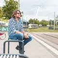 Ein Mann mittleren Alters mit Sonnenbrille sitzt an einem schönen Tag auf einer Bank an einer Bahn-Haltestelle.