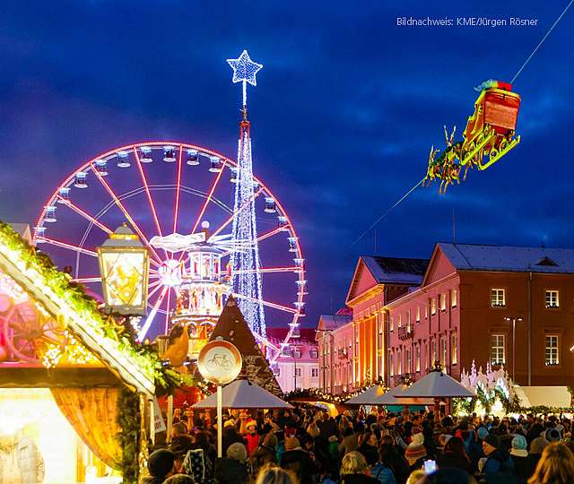 Weihnachtsmarkt Karlsruhe mit beleuchteten Gebäuden, Riesenrad und vielen Besuchern. Der fliegende Weihnachtmarkt fliegt mit seinen Rentieren über den weihnachtlich, geschmückten Platz.