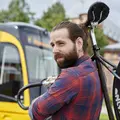 Ein junger Mann mit Bart hat ein Fahrrad über der Schulter. Er steht vor einer Bahn.