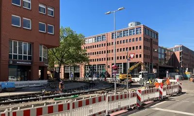 Das Foto zeigt die aktuellen Gleisbauarbeiten am Kronenplatz im Kruzungsbereich Kaiserstraße/Fritz-Erler-Straße. Zu sehen sind Bagger, Baumaschinen und Absperrbarken sowie Gebäudefassaden im Bildhintergrund.