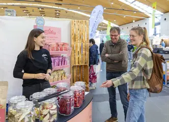 Zwei Messe-Besucher stehen an einem Messe-Stand bei der Fachmesse "expoSE" und "expoDirekt" und sprechen mit einer Frau, die den Messe-Stand betreut. Am Messestand stehen Einmachgläser mit Beeren und Früchten.