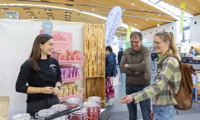 Zwei Messe-Besucher stehen an einem Messe-Stand bei der Fachmesse "expoSE" und "expoDirekt" und sprechen mit einer Frau, die den Messe-Stand betreut. Am Messestand stehen Einmachgläser mit Beeren und Früchten.