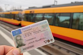 Eine Hand hält einen Führerschein im Scheckkarten-Format. Im Hintergrund fährt eine Straßenbahn.