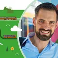 Ein Mann trägt eine Umhängetasche über der Schulter und lächelt in die Kamera. Er lehnt an einer Wand. Im Hintergrund ist eine Bahn zu sehen.
