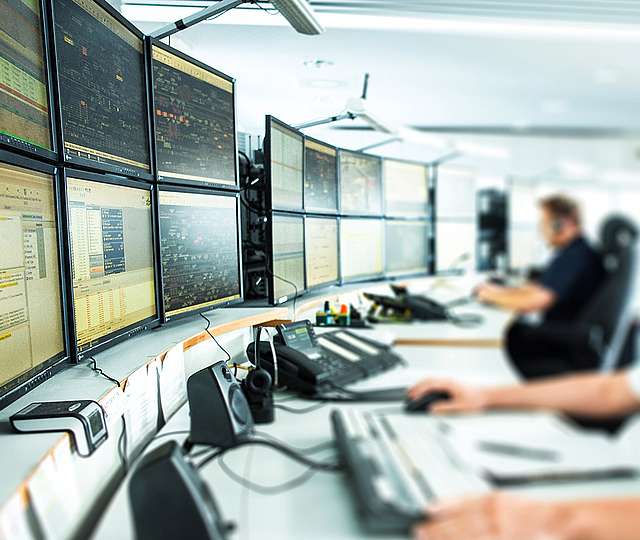 Zwei Männer überwachen und arbeiten an einer Wand mit zahlreichen Bildschirmen.