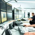 Zwei Männer überwachen und arbeiten an einer Wand mit zahlreichen Bildschirmen.