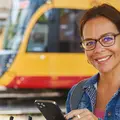 Eine Frau steht vor einem Voi-Roller und hält ihr Smartphone in der Hand. Sie lächelt dabei. Im Hintergrund ist eine Bahn zu sehen.