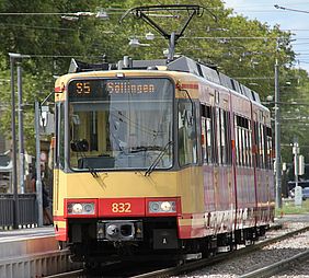 Eine rot-gelbe Stadtbahn der Linie s5 hält an einer Haltestelle. Im Hintergrund sind grüne Bäume zu sehen.