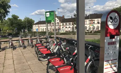 Räder des Bike-Sharing-Angebots kvv.netbike stehen an einer Ausleihstation auf dem Platz an der Reithalle in Rastatt. Im Hintergrund ist das Bahnhofsgebäude von Rastatt zu sehen