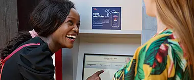 Eine junge Frau steht am Fahrkartenautomat und lacht. 