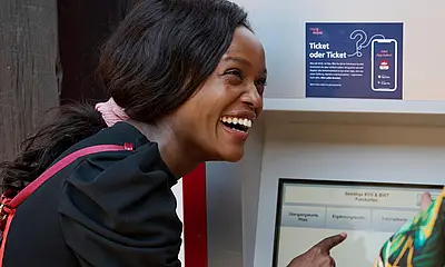 Eine junge Frau steht am Fahrkartenautomat und lacht. 