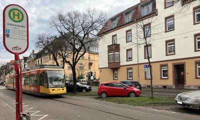 Das Foto zeigt die westliche Kriegsstraße im Bereich der Haltestelle Hübschstraße. Eine VBK-Trambahn fährt vorbei, daneben sind parkende Autos und Häuserfassaden zu sehen. Am linken Bildrand steht ein Haltestellenschild. 