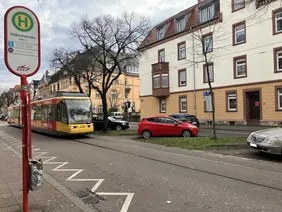 Das Foto zeigt die westliche Kriegsstraße im Bereich der Haltestelle Hübschstraße. Eine VBK-Trambahn fährt vorbei, daneben sind parkende Autos und Häuserfassaden zu sehen. Am linken Bildrand steht ein Haltestellenschild. 