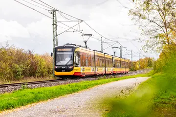 Eine Bahn der Linie S1/S11 fährt auf der Hardtbahn in Richtung Hochstetten. Hinter der Bahn sind eine Hecke und die Oberleitung zu sehen.