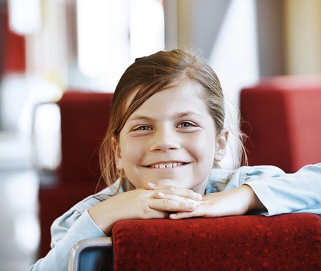 Ein Mädchen in der Bahn hat die Hände und das Kinn auf die Rückenlehne eines Sitzes gestützt und lächelt.