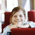 Ein Mädchen in der Bahn hat die Hände und das Kinn auf die Rückenlehne eines Sitzes gestützt und lächelt.