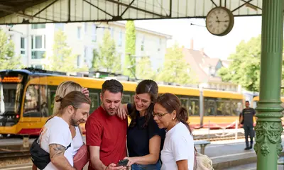 Fünf Menschen stehen in einem Halbkris und blicken gemeinsam auf ein Smartphone. Im Hintergrund ist eine Stadtbahn zu sehen.