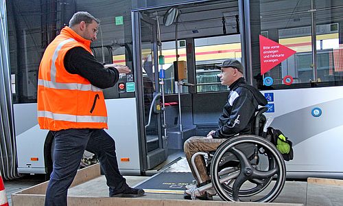 Ein Mitarbeiter der VBK mit Warnweste steht an der geöffneten Tür eines Busses und unterhält sich mit einem Fahrgast in einem Rollstuhl.