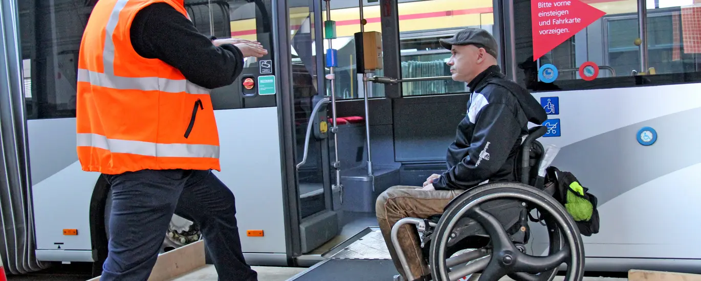 Ein Mitarbeiter der Verkehrsbetriebe Karlsruhe gibt einem Fahrgast, der in einem Rollstuhl sitzt, Tipps für den sicheren Ein- und Ausstieg bei einem Linienbus der VBK.